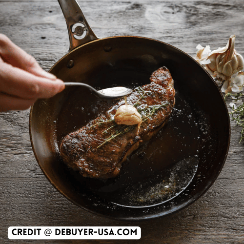a person holding a spoon over a piece of meat in a cast iron pan